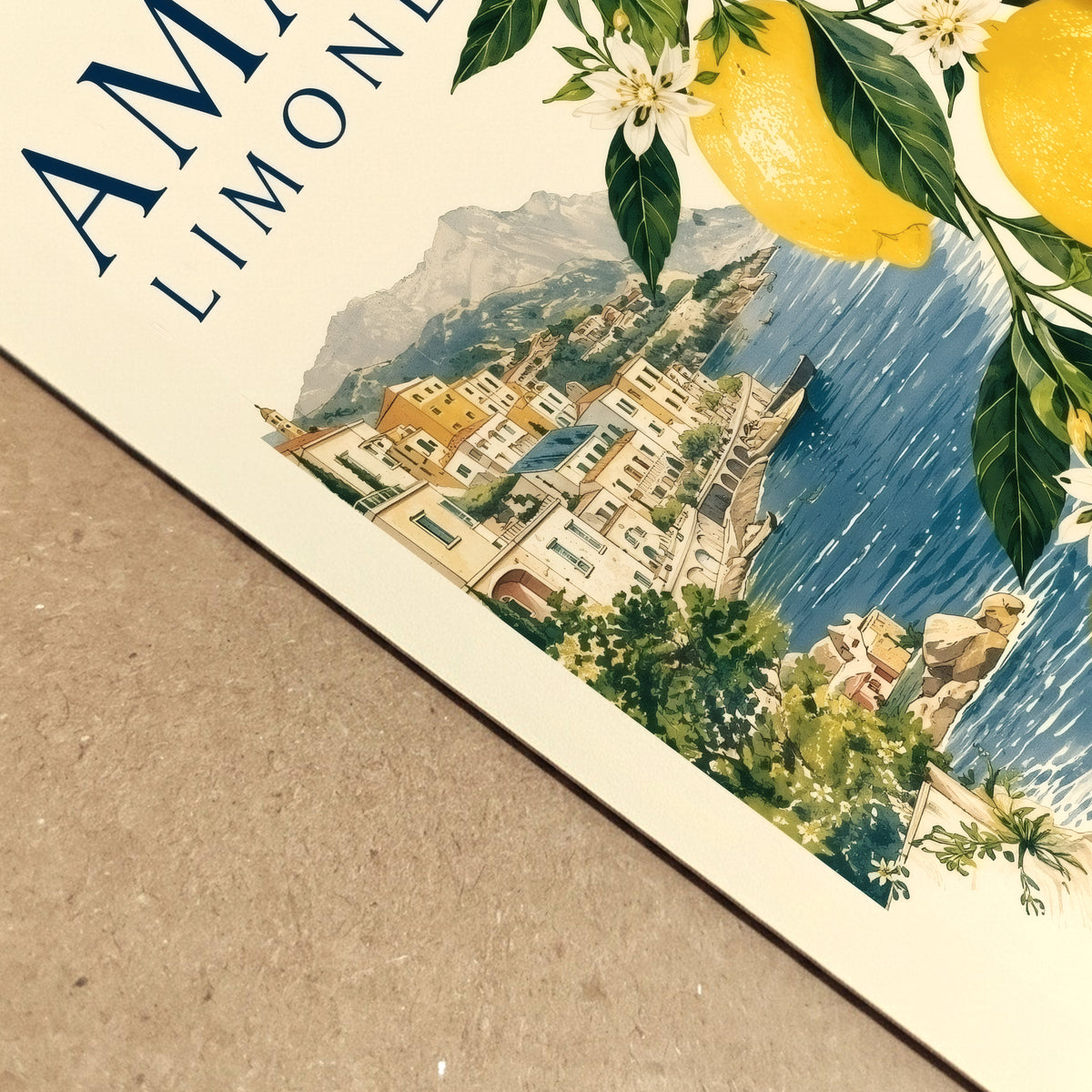 Close up of an Amalfi Coast lemon poster lying on a textured surface, showing the printed paper detail, yellow citrus fruit, green leaves, blue sea and Italian village artwork. A vintage style Mediterranean travel print with fresh summer colour and coastal character.