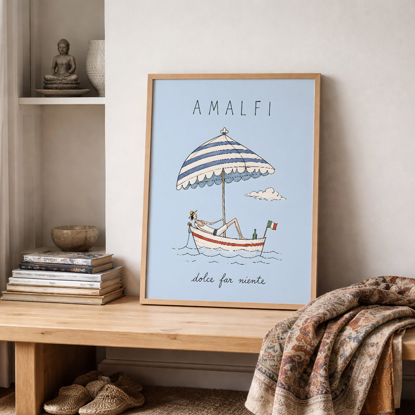 Amalfi boat poster resting on a wooden shelf in a neutral lived-in interior, surrounded by stacked books, pottery and folded textured fabrics in warm natural colours.