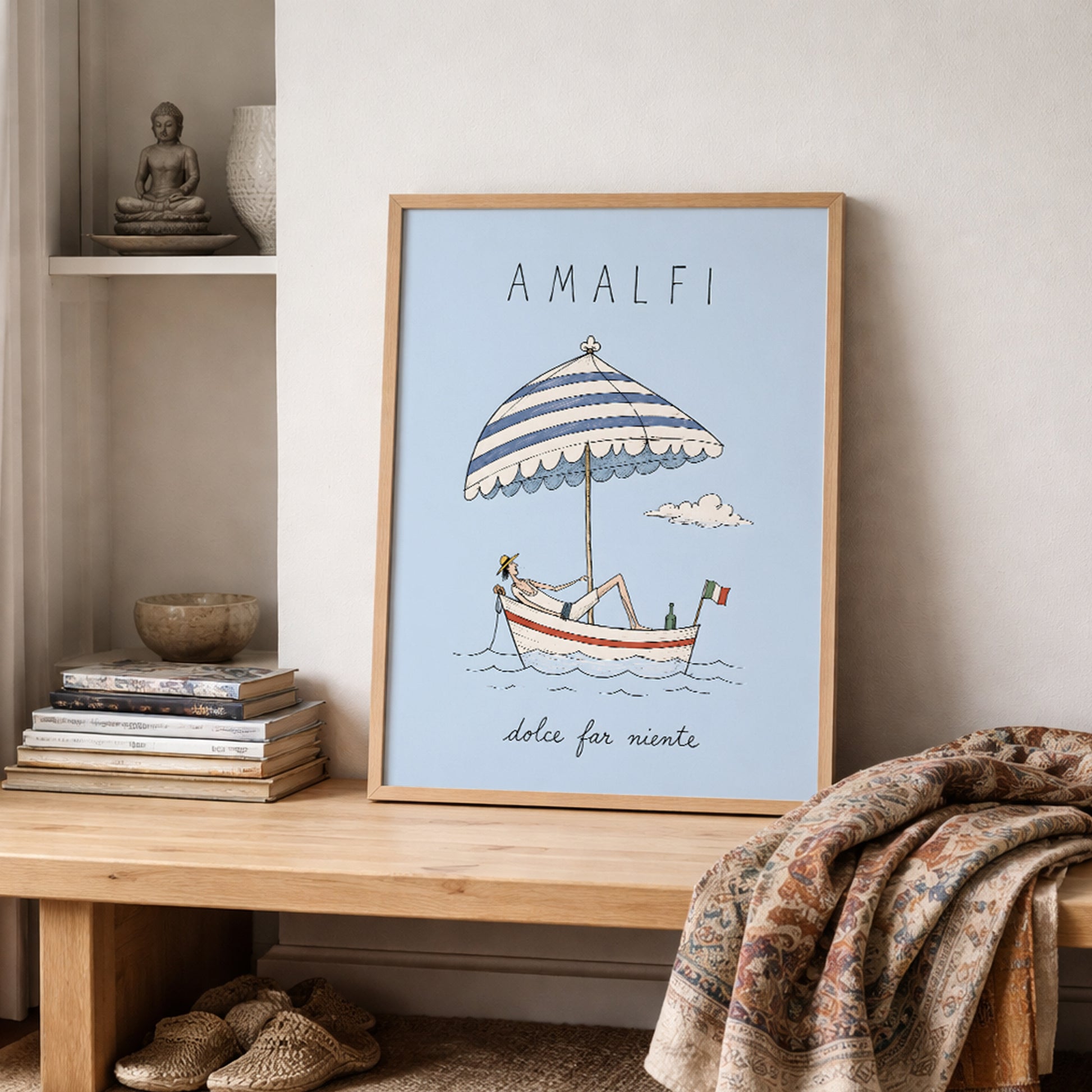 Amalfi boat poster resting on a wooden shelf in a neutral lived-in interior, surrounded by stacked books, pottery and folded textured fabrics in warm natural colours.