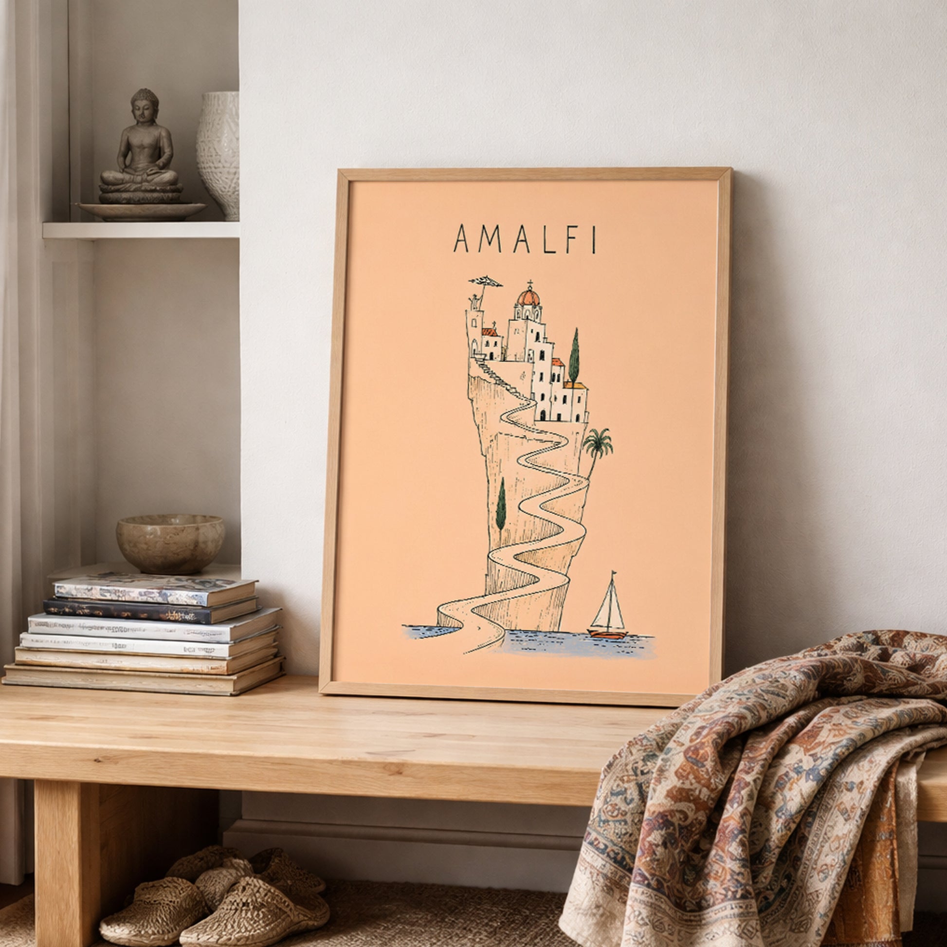 Unframed Amalfi Coast poster resting on a wooden shelf against a neutral wall, styled with stacked books, ceramics and a softly draped textured blanket.