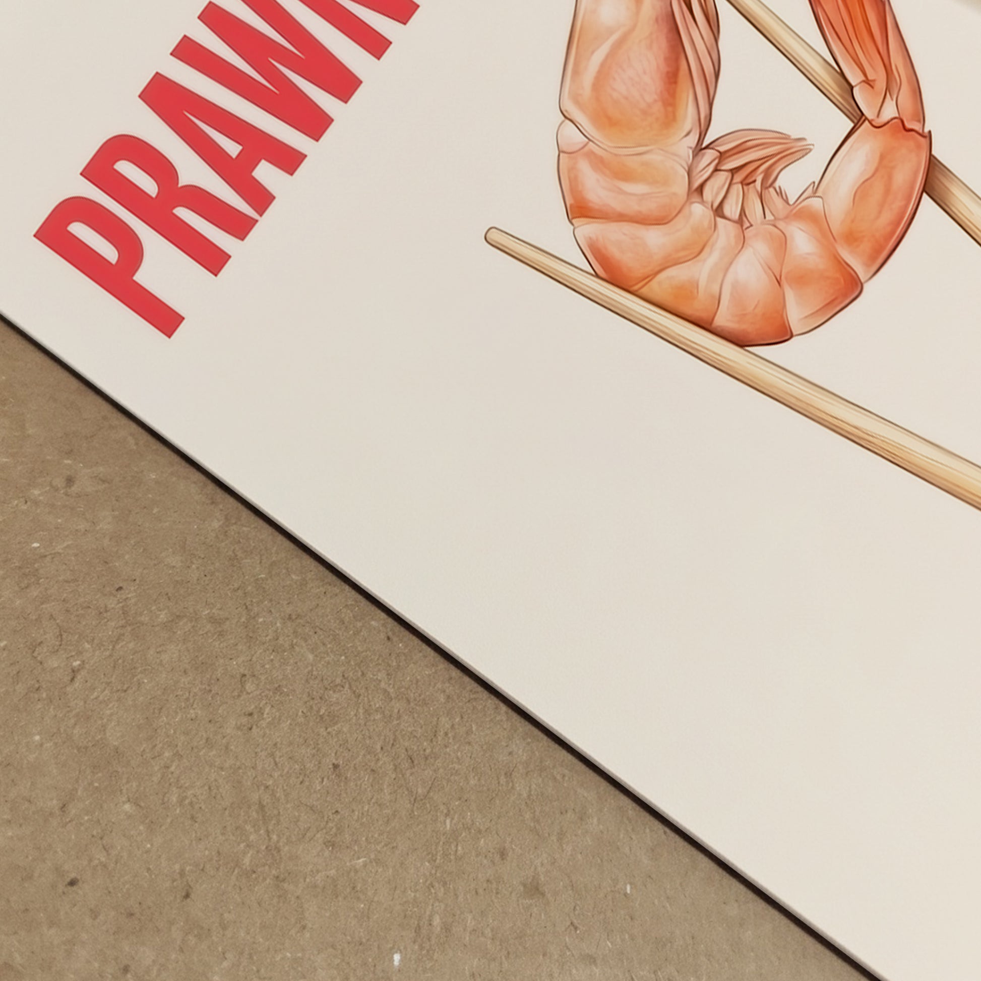 Angled close-up of the Prawnography shrimp food poster showing the top corner of the print, bold red PRAWNOGRAPHY text and part of the prawn held in chopsticks. The cream background and paper surface are visible in detail, highlighting the clean layout and modern graphic style. A funny food art print with Asian food inspiration, bold typography and a minimalist look for kitchen and dining wall decor.