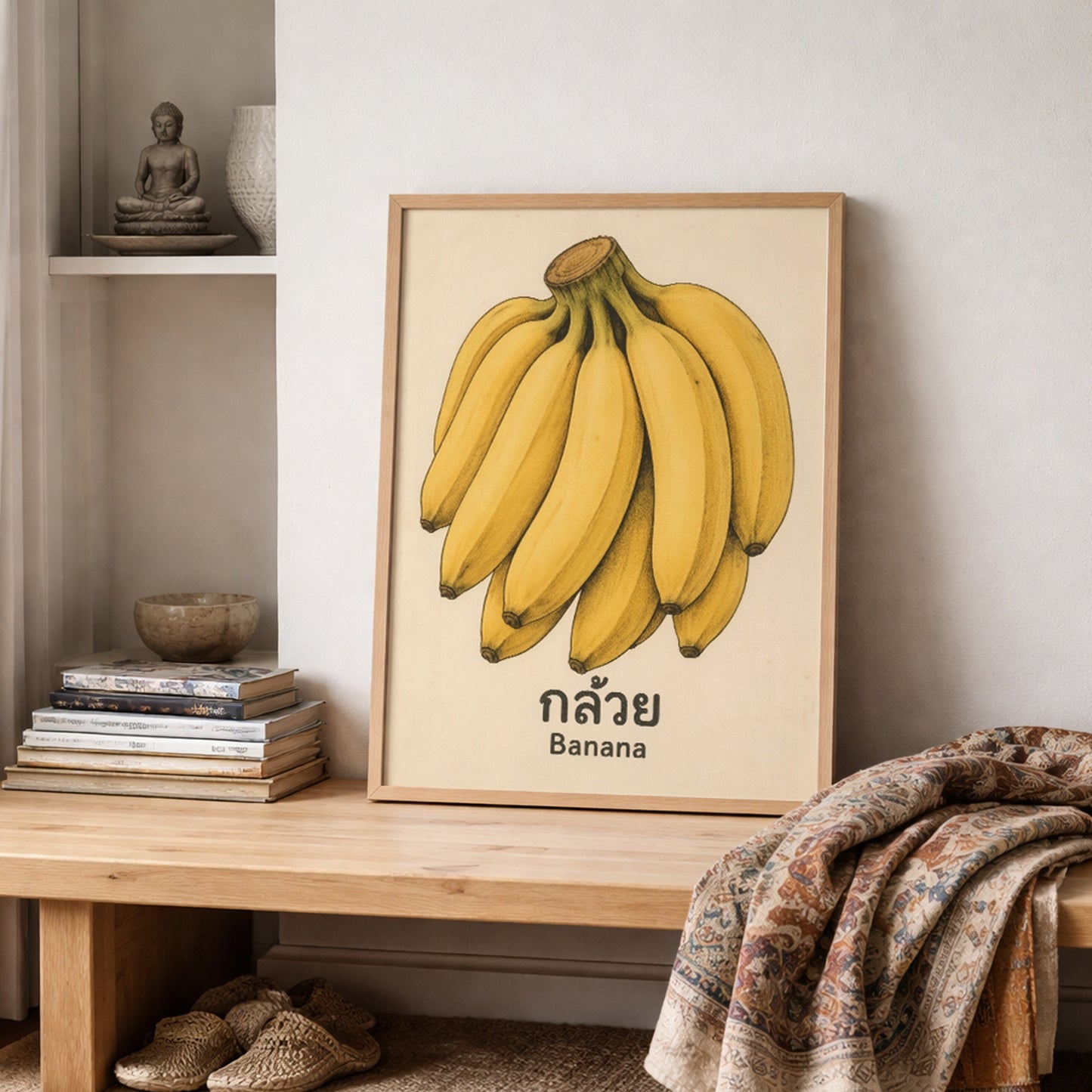 Framed vintage Thai banana poster styled on shelving in a calm interior with stacked books, neutral decor, and folded textiles draped nearby. The artwork features yellow bananas, Thai script, and an aged background that gives it a simple retro market print feel. This image shows how the piece fits into everyday home decor and supports searches for banana kitchen art, fruit poster, shelf wall art, and tropical room styling.