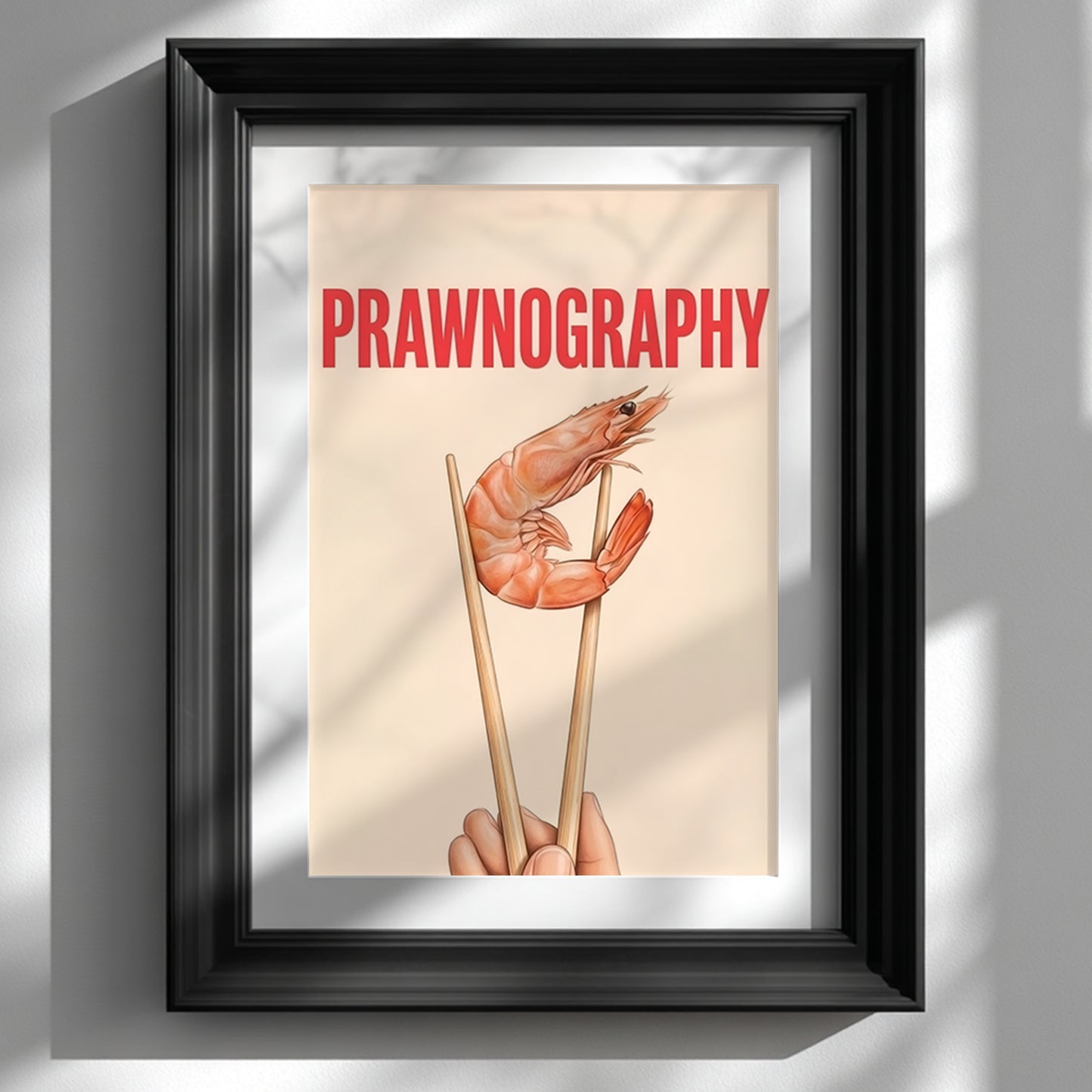 Framed Prawnography shrimp food poster shown on a white wall in a glossy black frame with a wide white mount. The artwork features a prawn held in chopsticks under bold red PRAWNOGRAPHY text on a soft cream background. Styled as funny kitchen wall art with modern typography, seafood wall decor and a clean minimalist look for dining rooms, home bars and contemporary interiors.
