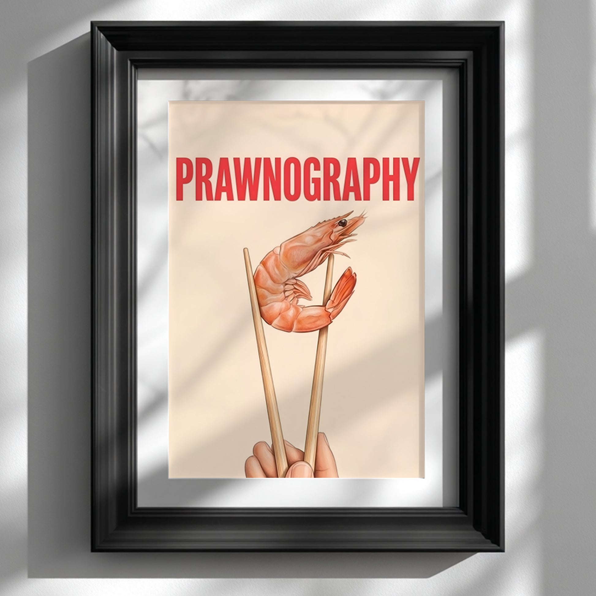 Framed Prawnography shrimp food poster shown on a white wall in a glossy black frame with a wide white mount. The artwork features a prawn held in chopsticks under bold red PRAWNOGRAPHY text on a soft cream background. Styled as funny kitchen wall art with modern typography, seafood wall decor and a clean minimalist look for dining rooms, home bars and contemporary interiors.