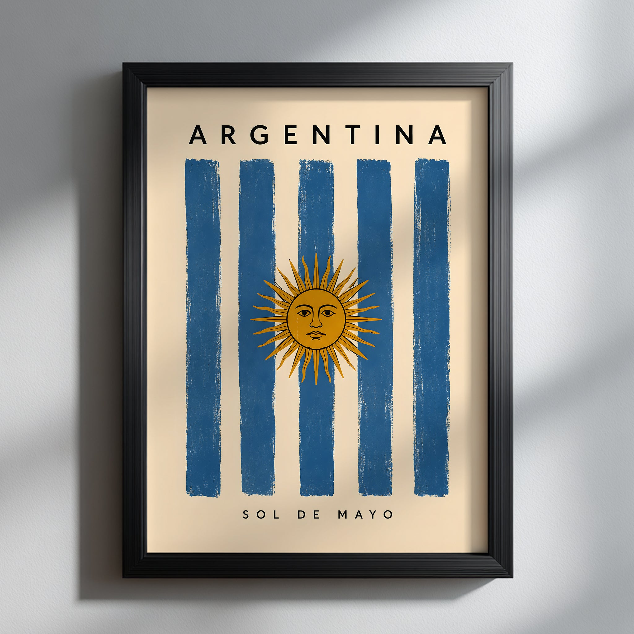Argentina flag poster presented in a slim black frame against a softly lit grey wall. The vertical artwork combines blue and cream stripes with the golden Sun of May, Argentina lettering and Sol de Mayo text. Strong natural shadows give this Argentine travel print a clean, contemporary appearance suited to neutral interiors.