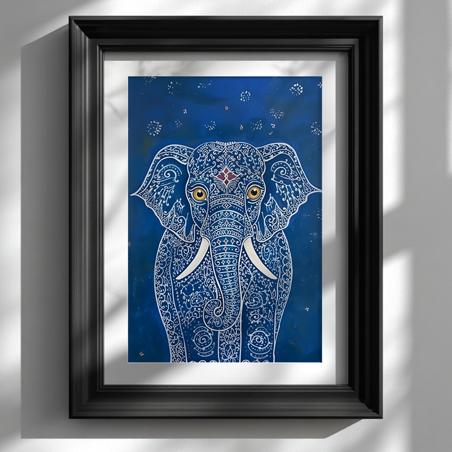 Black framed Blue Elegant Elephant Print displayed on a white wall with soft natural light and window shadows. The artwork shows a front-facing elephant with intricate white pattern lines, large gold eyes, white tusks and a small red forehead detail on a deep blue background. A clean interior mockup for blue elephant wall art, Indian elephant art and decorative animal print styling.