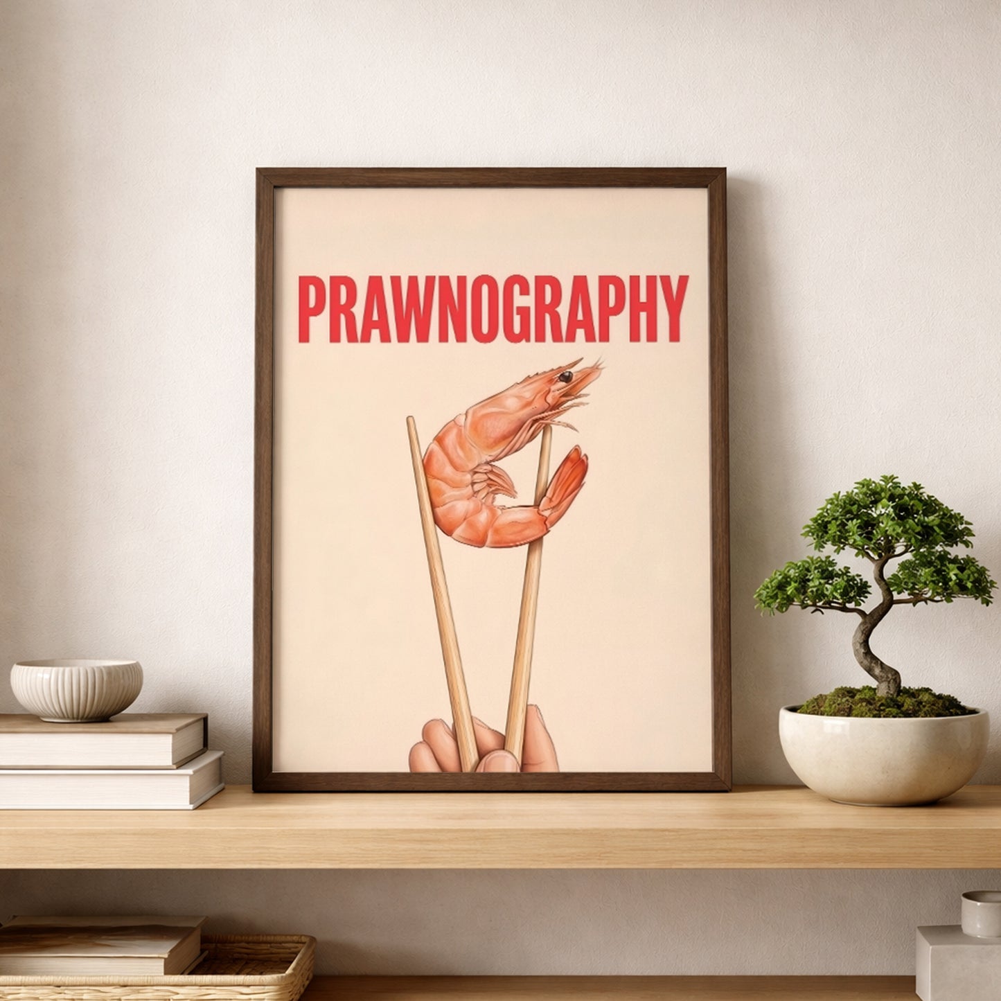Framed Prawnography shrimp food poster displayed on a wooden shelf beside stacked books and a small bonsai-style plant. The artwork shows a prawn held in chopsticks with bold red PRAWNOGRAPHY text on a soft cream background. Styled as modern kitchen wall art with a clean neutral palette, playful food humour and simple typography, ideal for dining rooms, shelves, home bars and minimal interiors.