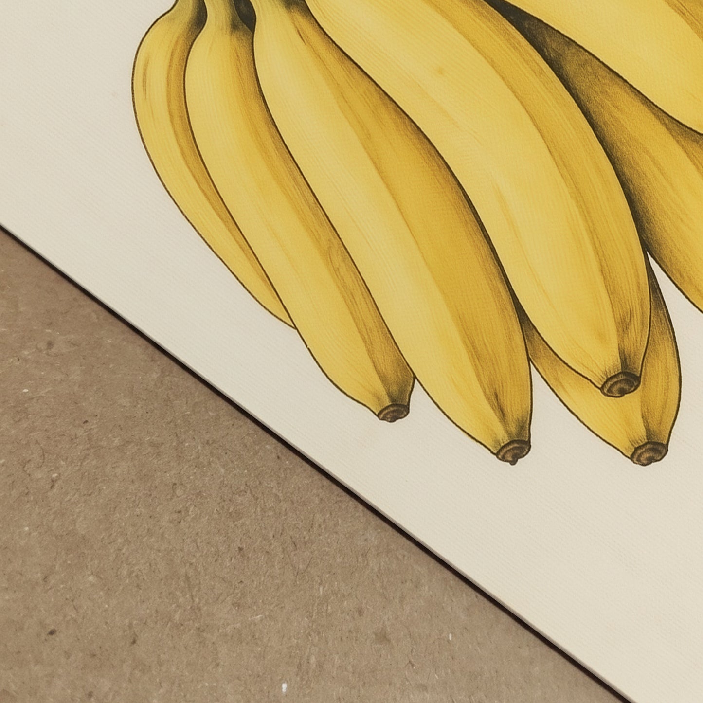 Close-up detail image of the banana print showing the lower part of the yellow bananas, the cream paper surface, and the visible paper edge resting on a textured backing board. The shot focuses on print finish, depth, and material quality rather than the full artwork. It is helpful for searches around banana art print detail, paper texture, premium poster quality, and botanical fruit wall art close-up views.