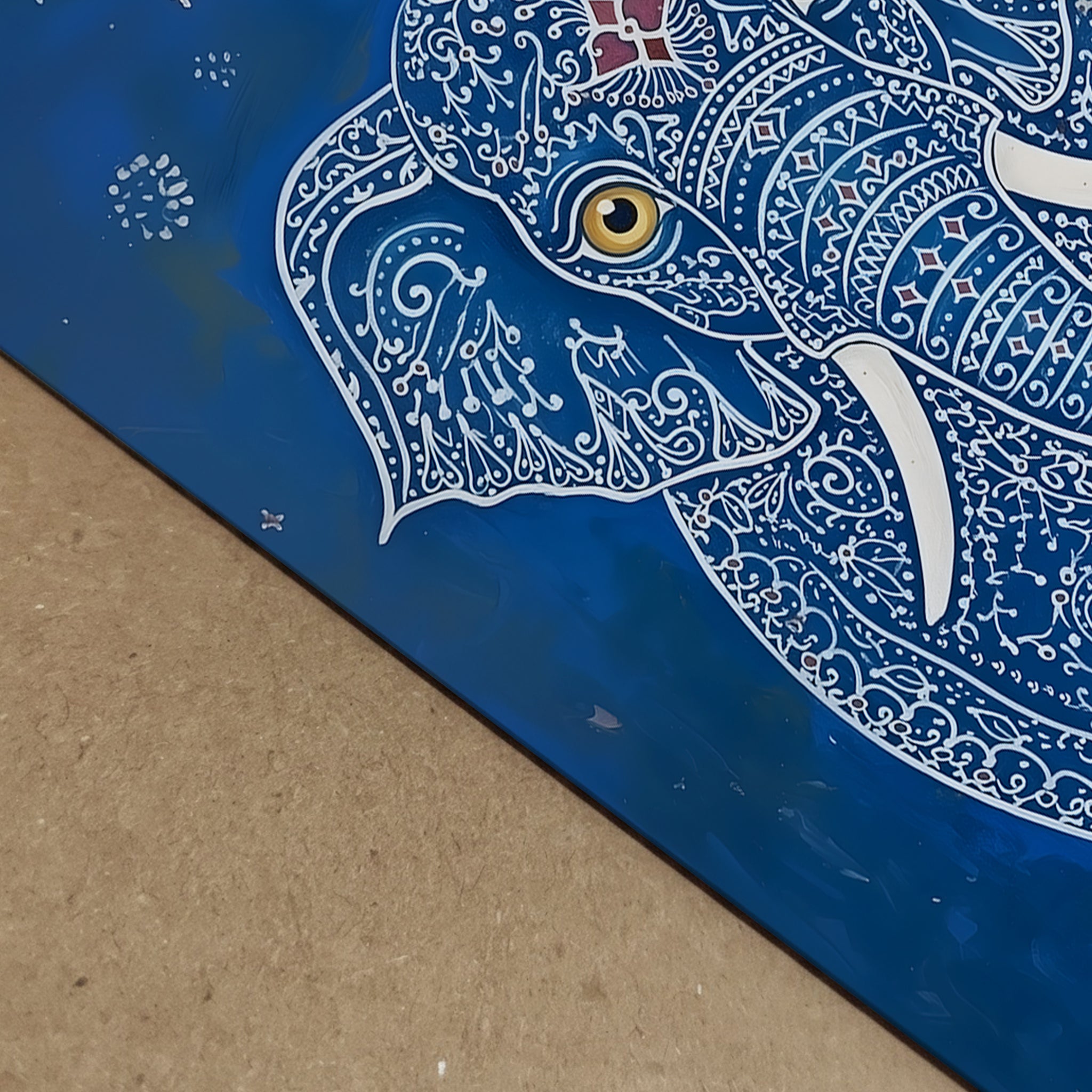 Close-up detail image of the Blue Elegant Elephant Print showing the artwork surface from an angled view. The visible section highlights the elephant’s gold eye, white tusk, white decorative linework and rich blue background, along with the edge of the print. A useful detail shot for blue elephant art print, Indian elephant artwork, ornate animal wall art and print quality presentation.