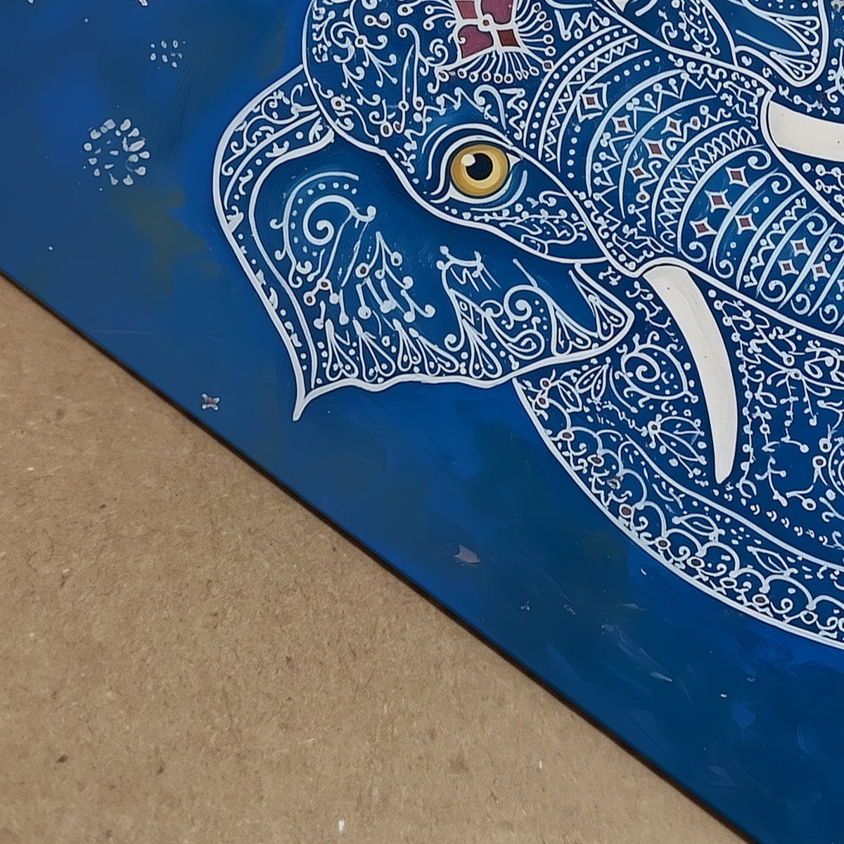 Close-up detail image of the Blue Elegant Elephant Print showing the artwork surface from an angled view. The visible section highlights the elephant’s gold eye, white tusk, white decorative linework and rich blue background, along with the edge of the print. A useful detail shot for blue elephant art print, Indian elephant artwork, ornate animal wall art and print quality presentation.