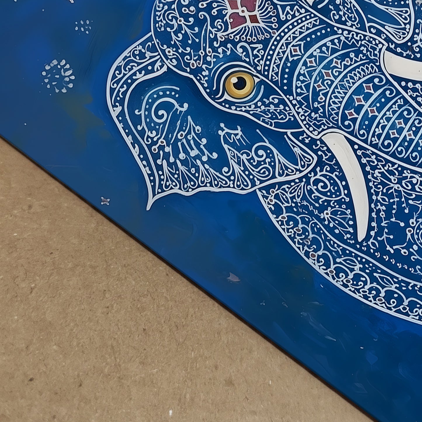 Close-up detail image of the Blue Elegant Elephant Print showing the artwork surface from an angled view. The visible section highlights the elephant’s gold eye, white tusk, white decorative linework and rich blue background, along with the edge of the print. A useful detail shot for blue elephant art print, Indian elephant artwork, ornate animal wall art and print quality presentation.