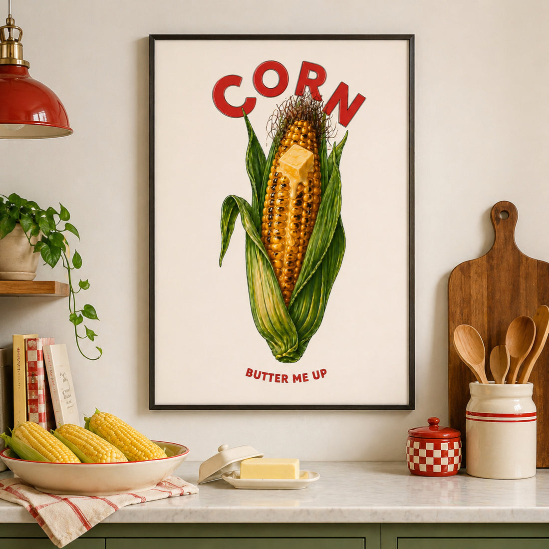 Framed corn butter poster displayed in a modern kitchen above a countertop with corn, butter, wooden utensils, green cabinets and red diner-style accents. The artwork shows a bright corn cob with green husks, melting butter, red CORN text and BUTTER ME UP below. A funny kitchen poster and food wall art print.
