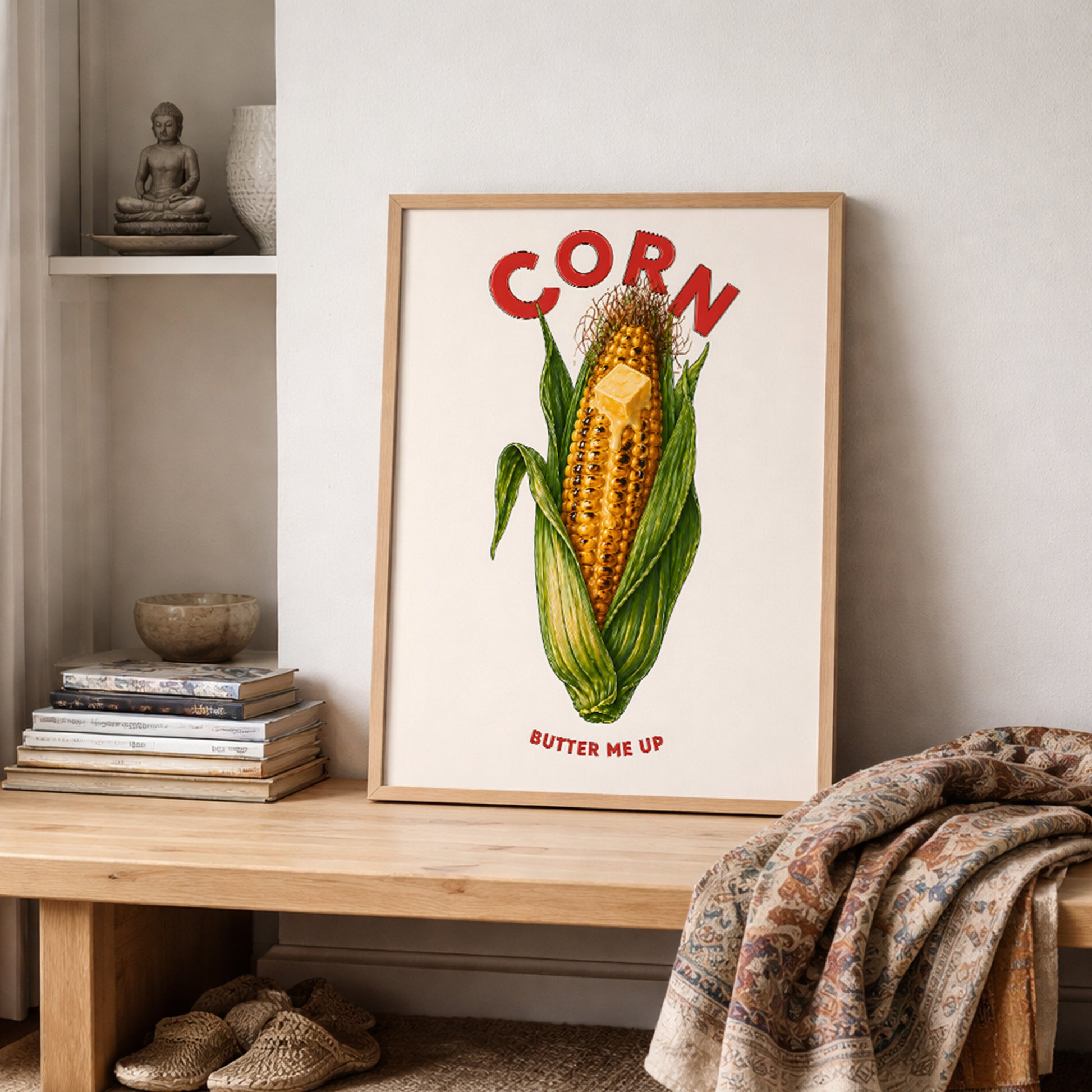 Corn butter poster styled on a shelf in a cosy room with books, soft fabric and neutral home decor. The artwork shows bright yellow corn, green husks, melting butter, red CORN text and BUTTER ME UP below. A fun food poster for kitchens, breakfast nooks, cafés and relaxed gallery walls.