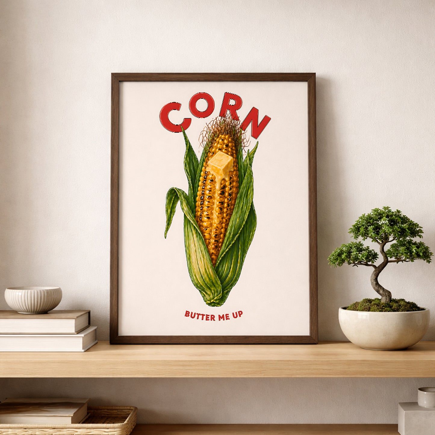 Corn butter poster displayed on a shelf beside a small bonsai-style plant in a calm modern room. The print shows a golden corn cob, green husks, melting butter and bold red lettering. A playful corn kitchen poster for shelves, dining spaces, cafés and simple food wall art styling.