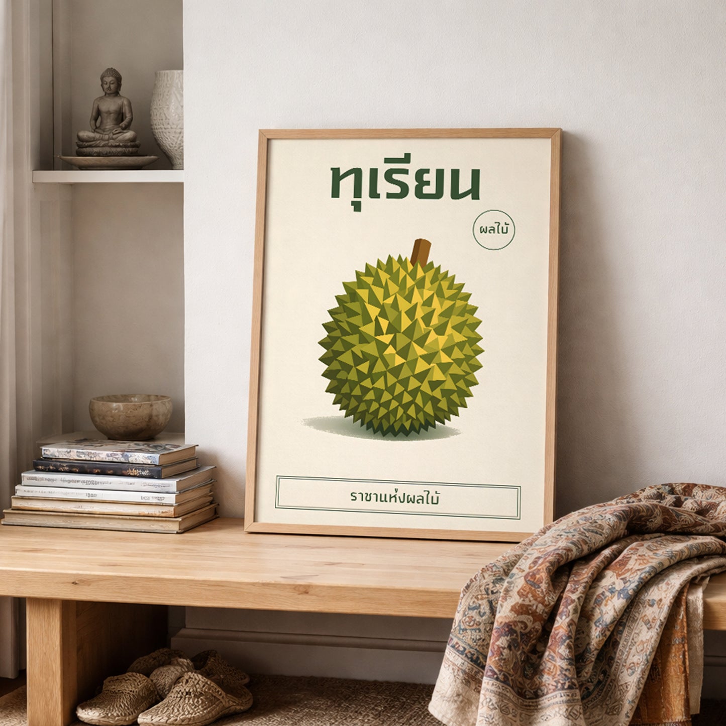 Botanical durian art print resting on a styled shelf with folded textiles and neutral home accessories. The framed illustration features a geometric green durian with Thai typography above. Warm lighting and layered fabrics create a cosy cultural aesthetic, ideal for kitchens, studios or relaxed contemporary living spaces.