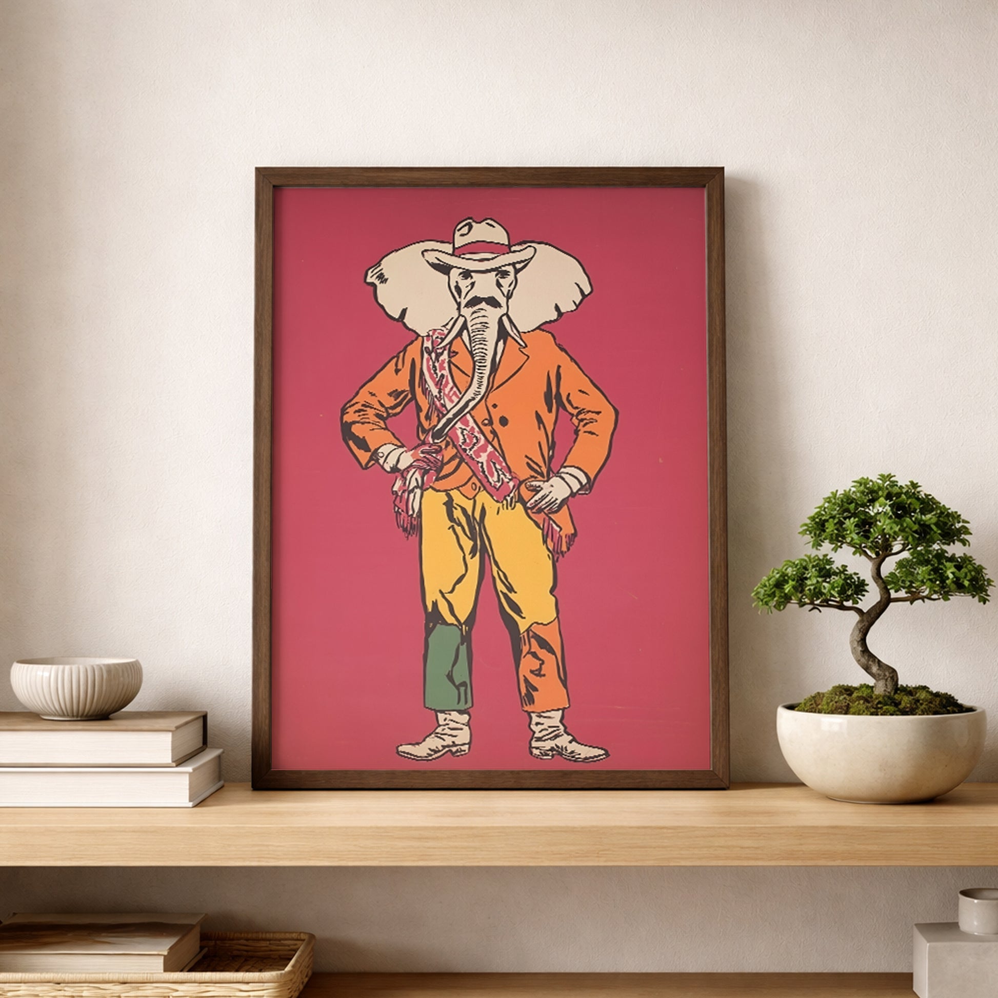 Elephant Cowboy poster leaning on a shelf in a clean modern room with stacked books and a bonsai plant. The print shows a playful elephant cowboy in a hat, orange jacket, yellow trousers and boots against a bold red background. Ideal as western wall art, cowboy poster or funny animal decor for minimalist homes, eclectic spaces, offices and modern gallery walls.