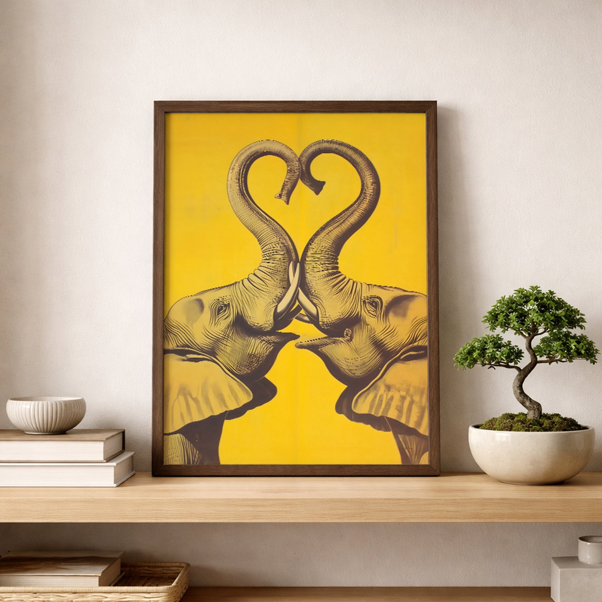 Framed Elephant Heart elephant print leaning on a shelf beside a small bonsai and minimal decor in a calm modern interior. The artwork shows two mirrored elephants with raised trunks creating a heart on a bright yellow background. Clean lifestyle image for elephant wall art, yellow elephant poster, framed elephant art, modern elephant decor, and asian-inspired wall art.