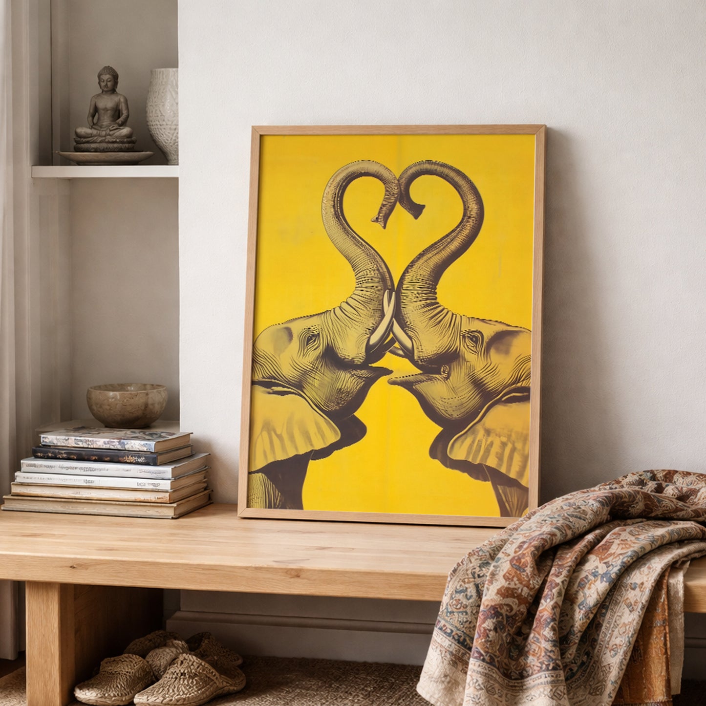 Framed Elephant Heart elephant wall art displayed on a shelf with stacked books, pottery, and textured fabric in a soft neutral room. The print features two mirrored elephants facing each other with trunks forming a heart over a bold yellow background. Styled image for elephant poster, elephant print, framed elephant wall decor, romantic elephant art, and cultural home decor.