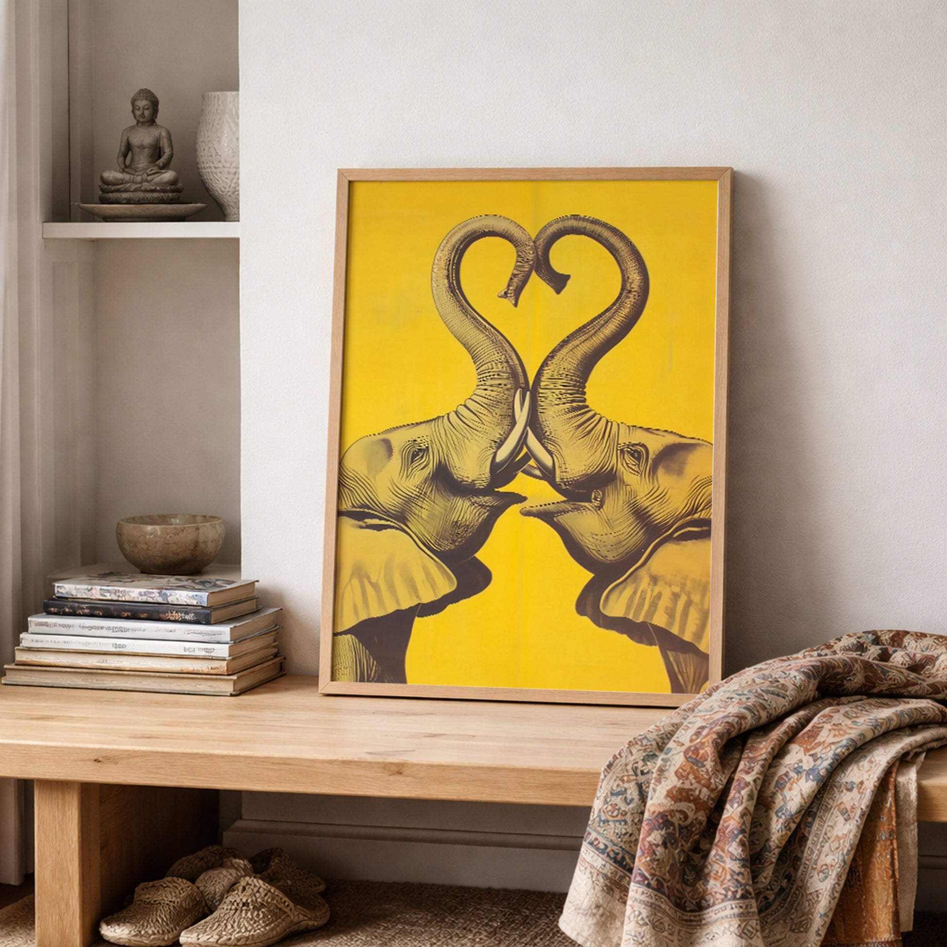 Framed Elephant Heart elephant wall art displayed on a shelf with stacked books, pottery, and textured fabric in a soft neutral room. The print features two mirrored elephants facing each other with trunks forming a heart over a bold yellow background. Styled image for elephant poster, elephant print, framed elephant wall decor, romantic elephant art, and cultural home decor.