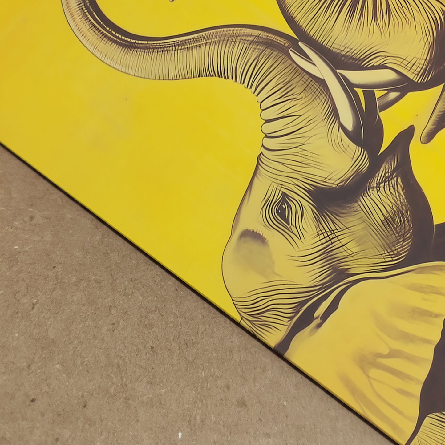 Close-up detail shot of the Elephant Heart yellow elephant print laid on a textured surface, showing the fine linework, paper edge, and part of one elephant with the curved trunk above. The bold yellow background and dark illustration give a sharp modern finish. Detail image for elephant art print, yellow elephant poster, premium paper print, and elephant wall decor listings.