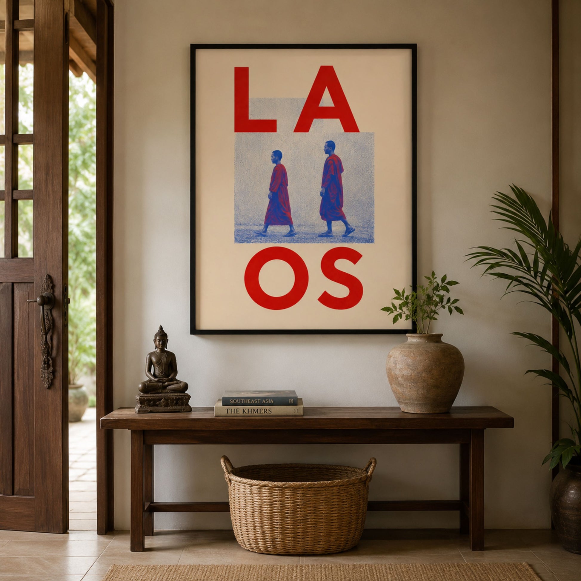 Framed Laos monk art displayed above a dark wooden console in a warm, lived-in interior. The red, blue and cream travel print is styled with a leafy plant, woven basket and simple decorative objects.