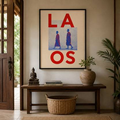 Framed Laos monk art displayed above a dark wooden console in a warm, lived-in interior. The red, blue and cream travel print is styled with a leafy plant, woven basket and simple decorative objects.