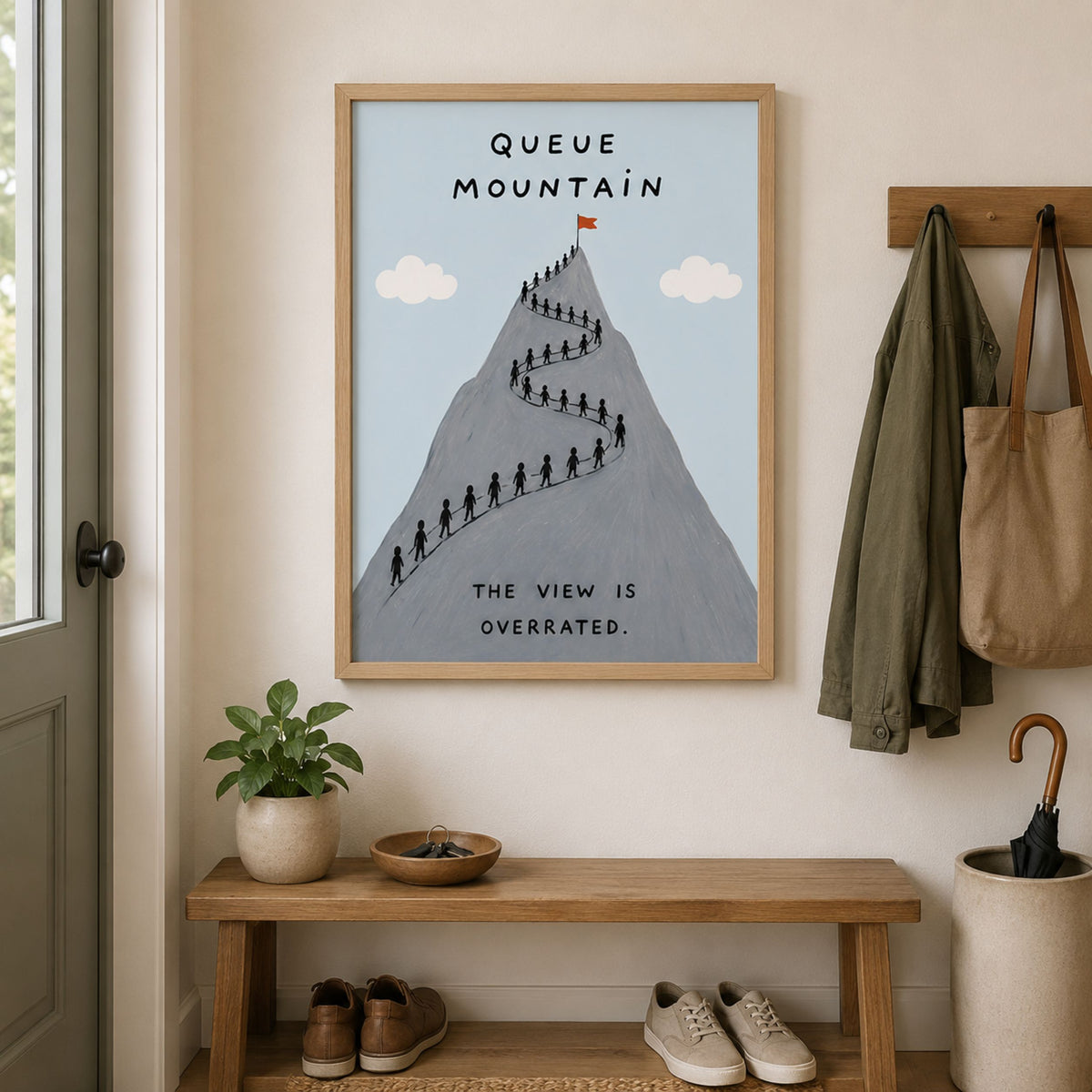 Framed Queue Mountain poster displayed above a wooden hallway bench with simple home decor, coats and plants. The grey mountain artwork shows tiny hikers queuing along a winding trail under a pale blue sky, with the text “Queue Mountain” and “The view is overrated” for funny hiking wall art.