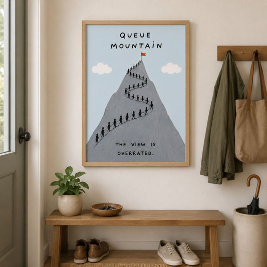 Framed Queue Mountain poster displayed above a wooden hallway bench with simple home decor, coats and plants. The grey mountain artwork shows tiny hikers queuing along a winding trail under a pale blue sky, with the text “Queue Mountain” and “The view is overrated” for funny hiking wall art.