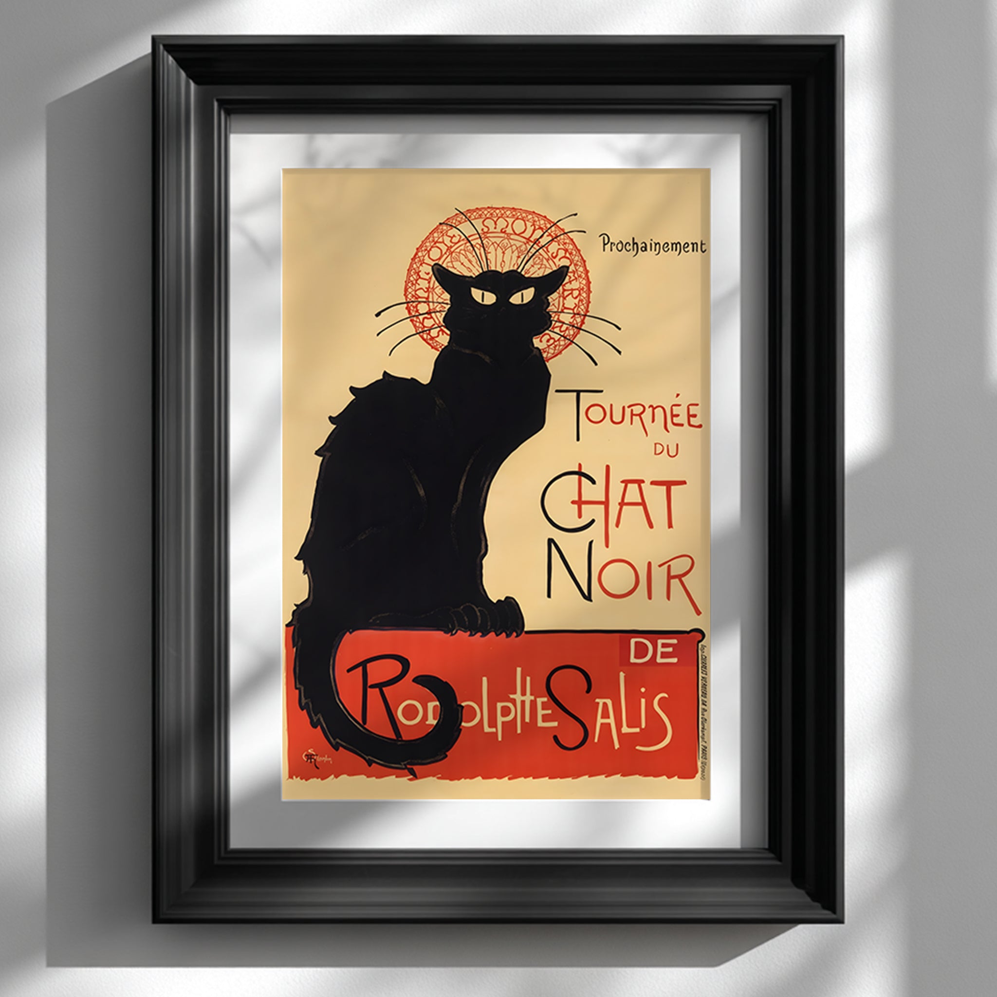 Framed Tournée du Chat Noir print by Théophile Steinlen displayed against a clean white wall in a classic black frame. The French Art Nouveau poster features the iconic black cat silhouette with yellow eyes and a red circular motif, alongside bold vintage typography. This Paris cabaret theatre poster adds striking black and red contrast to minimalist interiors and modern gallery wall decor.