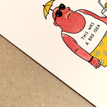 Close-up view of a funny travel poster printed on paper, showing the sunburnt cartoon character at an angle with sunglasses, a tiny umbrella hat, yellow flower shorts and a drink. The simple black linework and dry holiday joke make it a quirky summer travel print with a handmade cartoon feel.