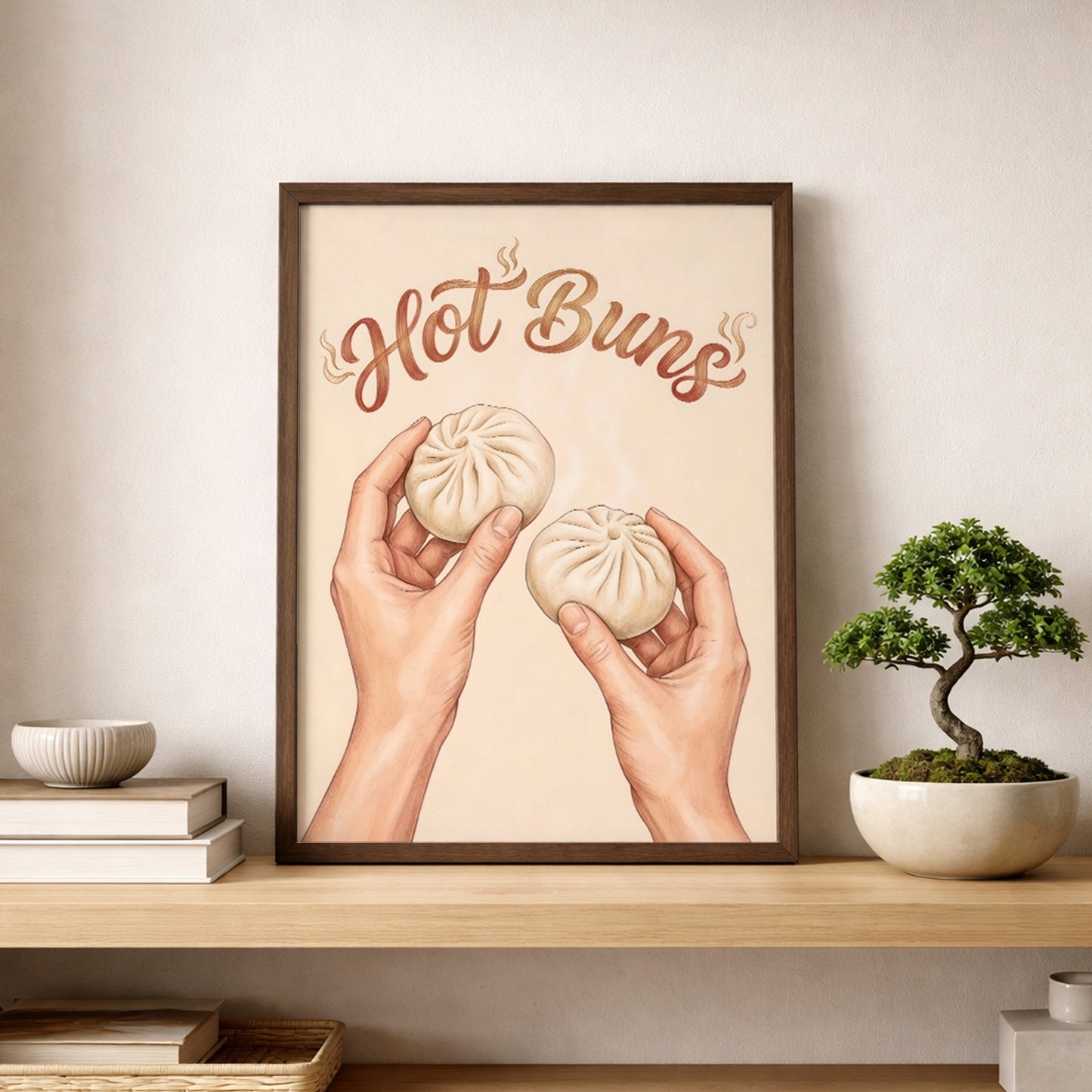 Hot Buns food print displayed on a shelf in a light wood frame beside a small bonsai-style plant and neutral home decor. The artwork features steamed buns held in two hands with brown script lettering on a soft beige background. The styling gives a clean modern kitchen feel and shows how the print can work as funny kitchen wall art, foodie decor, or simple asian food wall art.