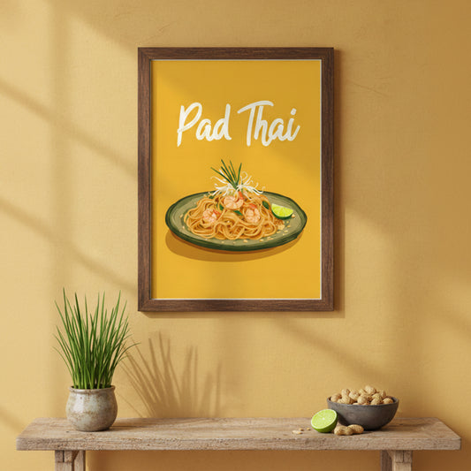 Framed kitchen poster on a warm yellow wall featuring an illustration of a plate of Pad Thai on a green dish with prawns, noodles, bean sprouts, chives and a lime wedge. The words “Pad Thai” appear in white script at the top against a mustard-yellow background. Below the frame is a wooden shelf holding a small potted grass plant, a bowl of peanuts and a halved lime.