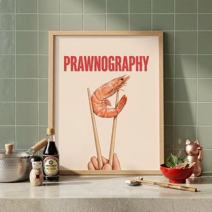 Prawnography shrimp food poster styled on a kitchen mantle or shelf against green tiled walls. The print sits in a light wood frame and shows a prawn held in chopsticks beneath bold red PRAWNOGRAPHY text on a cream background. Surrounded by kitchen items, bottles and bowls, it reads as funny kitchen wall art, seafood decor and modern typography art for cooking spaces, dining areas and shelves.