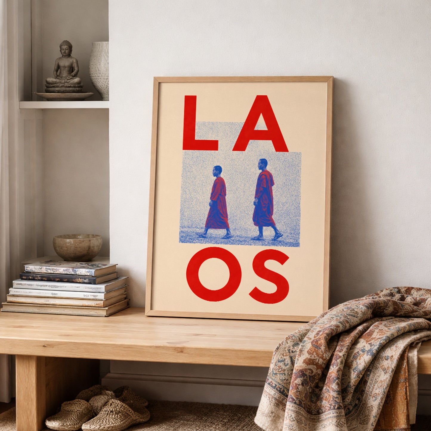 Wooden-framed Laos monk print resting on a shelf beside stacked books, pottery and folded patterned textiles. The red, blue and cream artwork adds a cultural travel theme to a neutral interior.