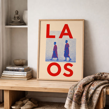 Wooden-framed Laos monk print resting on a shelf beside stacked books, pottery and folded patterned textiles. The red, blue and cream artwork adds a cultural travel theme to a neutral interior.
