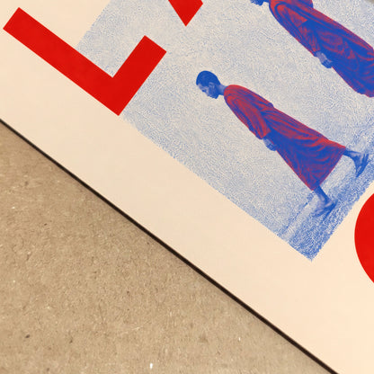 Close-up of the Laos monk art print lying on a textured surface, showing the cream paper, blue halftone pattern, red lettering and detailed figures of two Buddhist monks in red robes.
