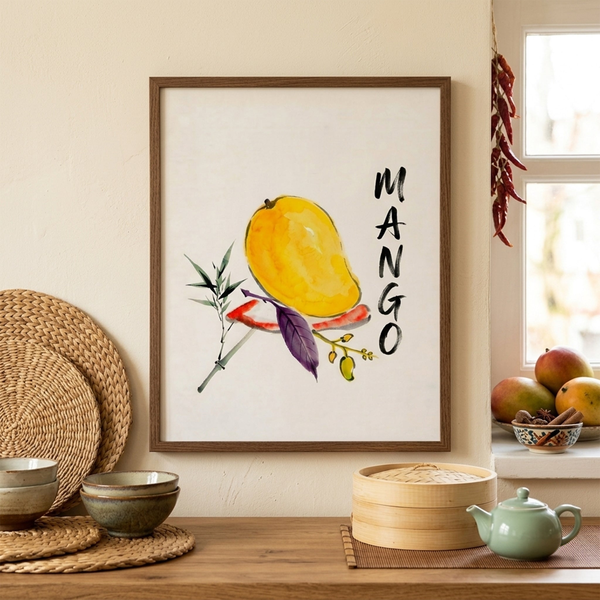 Mango wall art displayed in a light wood frame on a kitchen counter, styled with woven decor, ceramics and fresh fruit. The print shows a yellow mango with loose watercolour brushwork, green and purple leaves and bold vertical MANGO lettering on a white background. Warm neutral styling and natural textures show how the tropical fruit print works as kitchen wall art, fruit decor or a simple botanical print in everyday interiors.