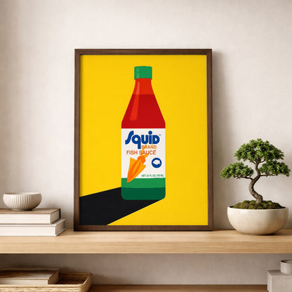 Squid Fish Sauce pop art poster framed and displayed on a shelf against a neutral wall, styled with stacked books and a small bonsai plant. The print features a graphic Thai fish sauce bottle on a vivid yellow background, creating a strong focal point. This mockup presents the artwork as modern kitchen wall art, Asian food poster decor and colourful pop art for minimalist shelves, home bars or dining spaces.