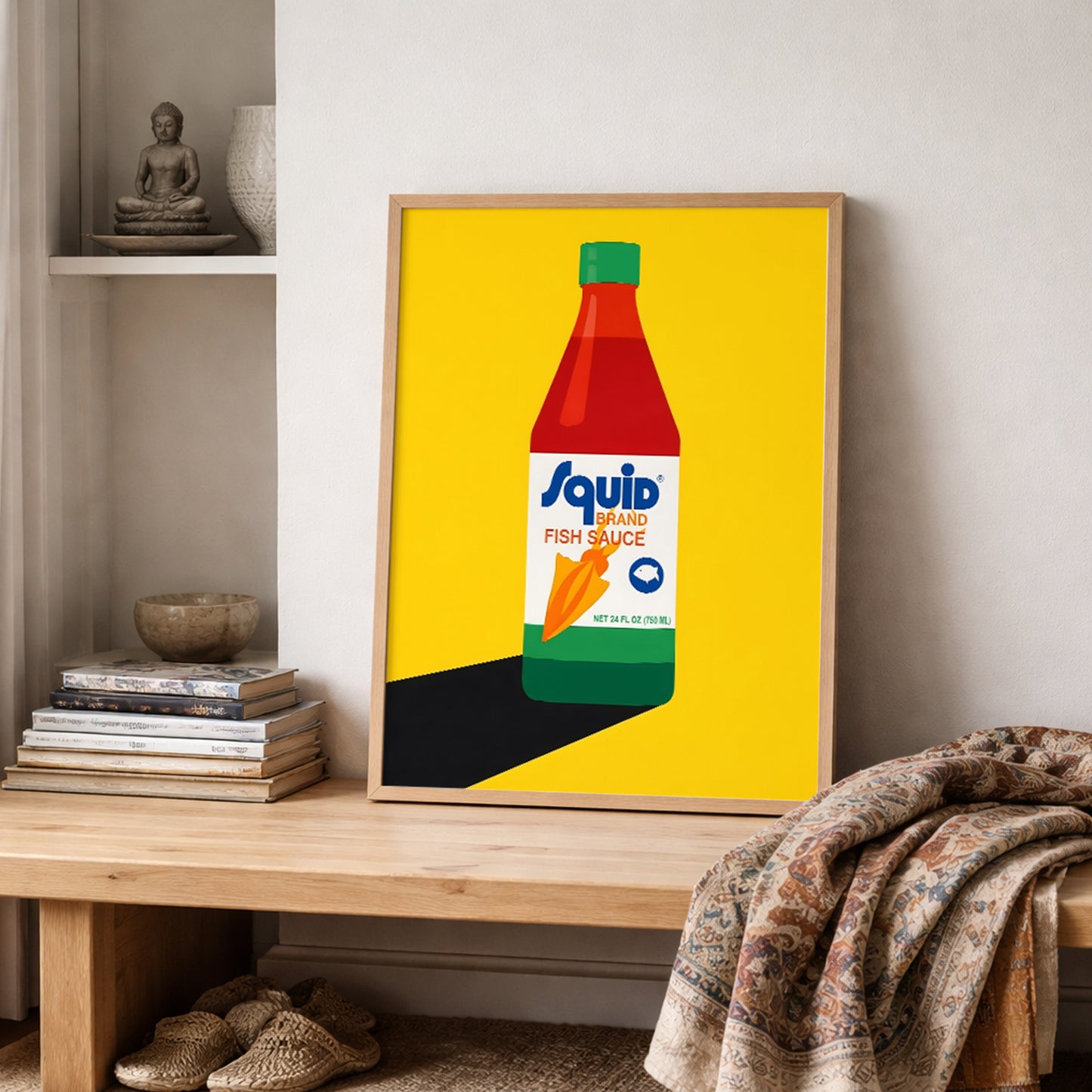 Framed Squid Fish Sauce pop art poster shown on a shelf in a cosy interior with books and folded textured fabric nearby. The artwork features a bold Thai fish sauce bottle illustration set against a bright yellow background with clean graphic shapes. This lifestyle image positions the print as quirky kitchen wall art, food-themed pop art and modern Asian decor that adds colour and character to shelves, dining rooms or gallery walls.