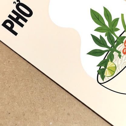 Close-up of a Vietnamese pho art print showing bold PHỞ lettering, green herbs and part of the illustrated noodle bowl. The cream printed surface rests against a natural brown textured background.