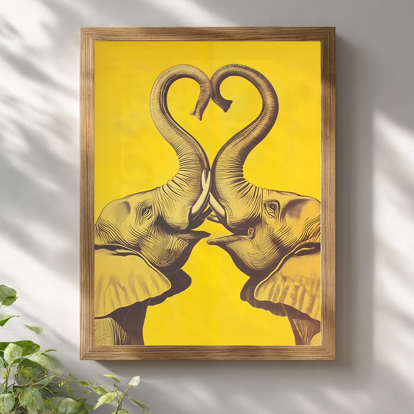 Wooden framed Elephant Heart elephant poster hung on a white wall with soft natural light and plant styling. The design shows two mirrored elephants facing inward, their trunks lifted into a heart shape in dark linework on a bold yellow background. Bright mockup for elephant wall art, elephant print, yellow elephant wall decor, framed elephant art, and modern cultural interiors.