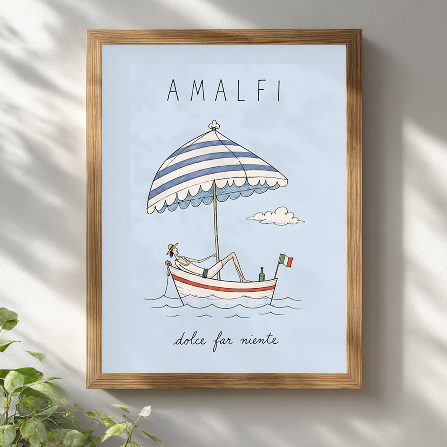 Amalfi Italy boat poster displayed in a light wooden frame on a white wall, with soft leafy shadows creating a relaxed summer setting around the pale blue travel artwork.