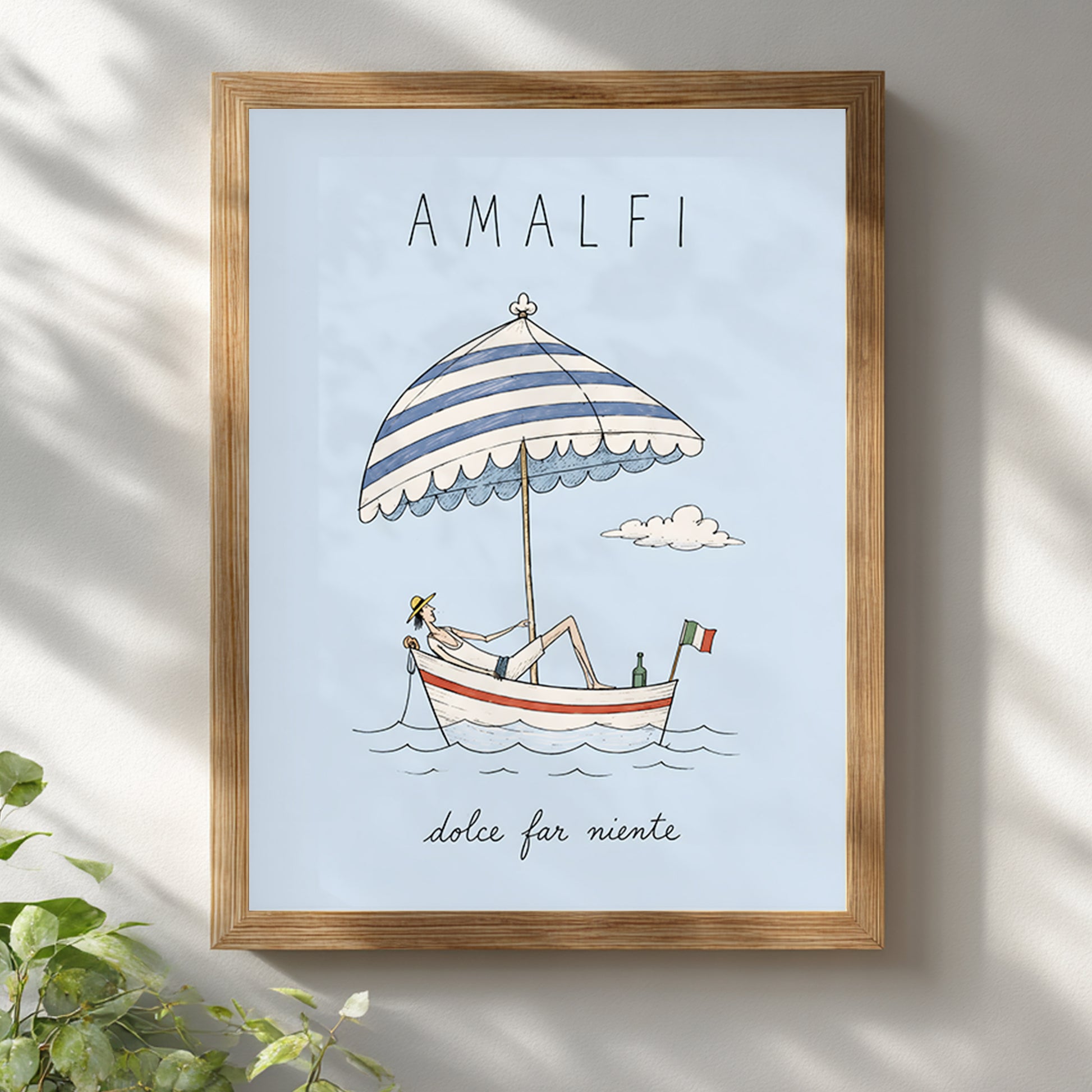 Amalfi Italy boat poster displayed in a light wooden frame on a white wall, with soft leafy shadows creating a relaxed summer setting around the pale blue travel artwork.