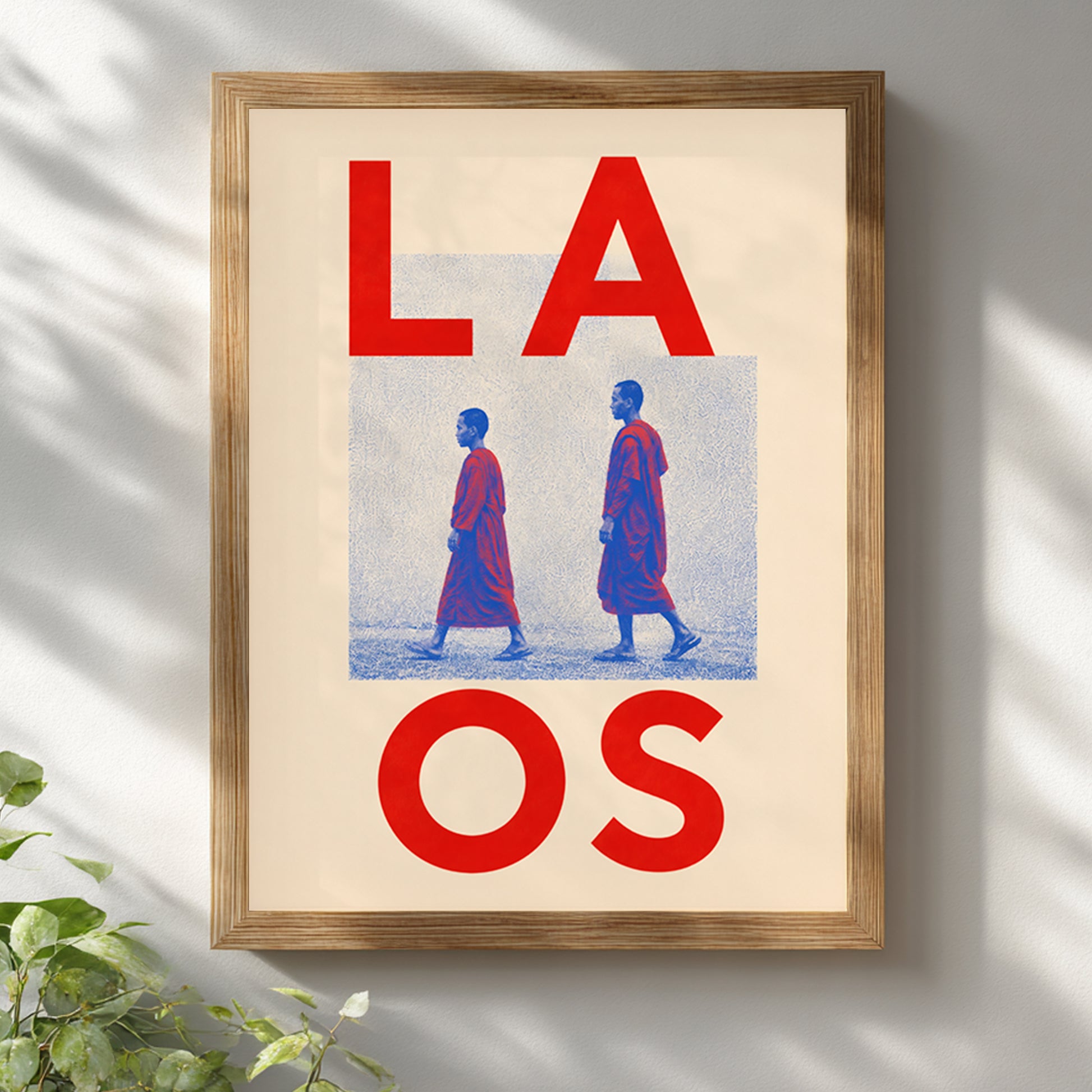 Wooden-framed Laos Buddhist monk wall art displayed on a white wall with soft natural shadows. Red LAOS typography, blue halftone figures and a cream background create a clean travel-inspired look.