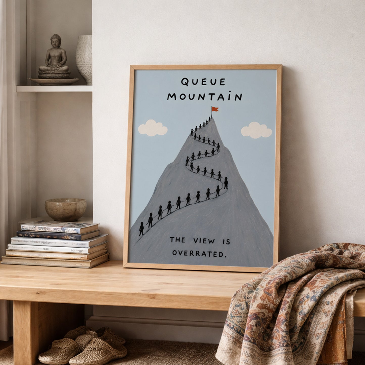 Wooden framed Queue Mountain poster styled on a shelf beside neutral home decor and soft fabric. The artwork shows a grey mountain with a long queue of tiny hikers, pale blue sky, cream clouds and the words “Queue Mountain” and “The view is overrated”, suited to funny hiking and travel wall decor.