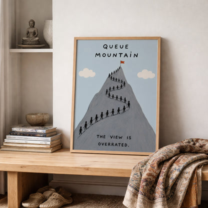 Wooden framed Queue Mountain poster styled on a shelf beside neutral home decor and soft fabric. The artwork shows a grey mountain with a long queue of tiny hikers, pale blue sky, cream clouds and the words “Queue Mountain” and “The view is overrated”, suited to funny hiking and travel wall decor.