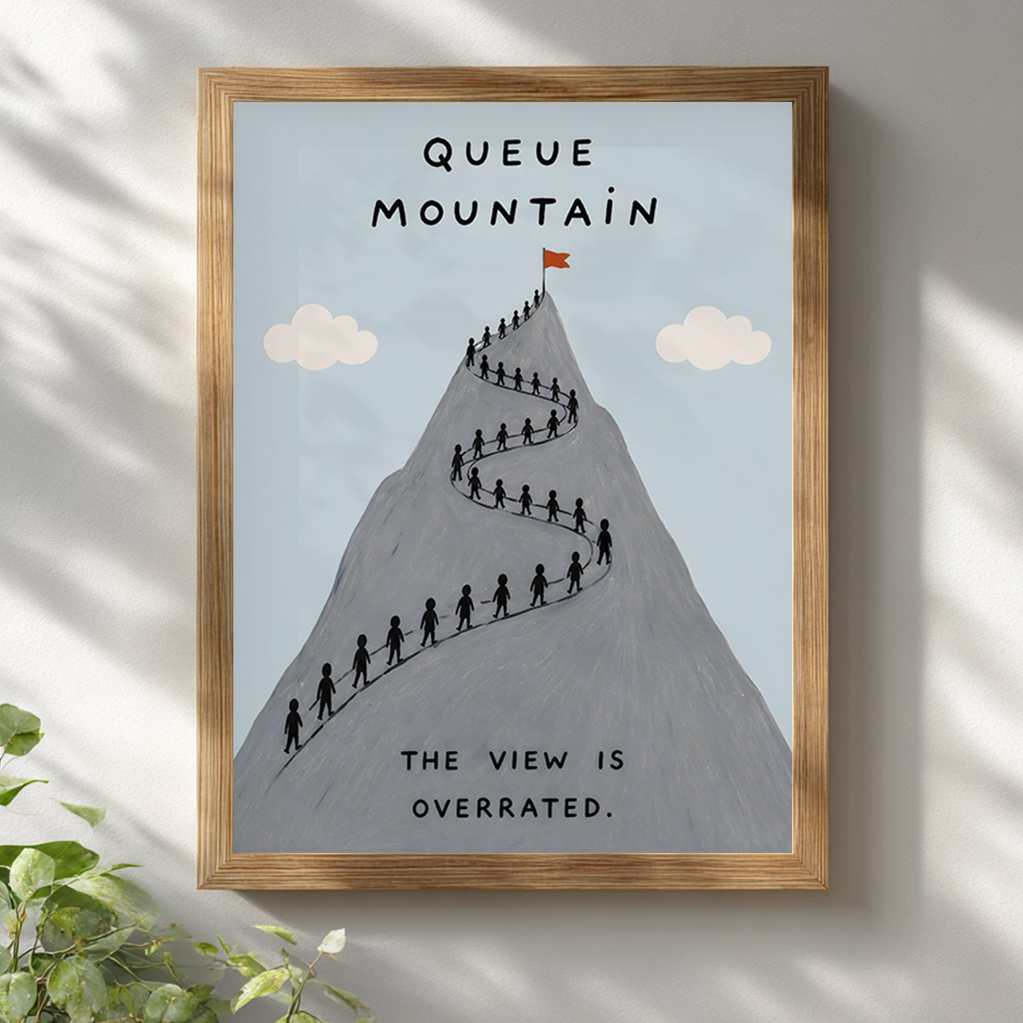 Wooden framed Queue Mountain poster hanging on a bright white wall with soft natural shadows. The artwork shows a grey mountain filled with tiny hikers queuing along a winding route, pale blue sky, cream clouds and the words “Queue Mountain” and “The view is overrated”, ideal for funny hiking wall art.